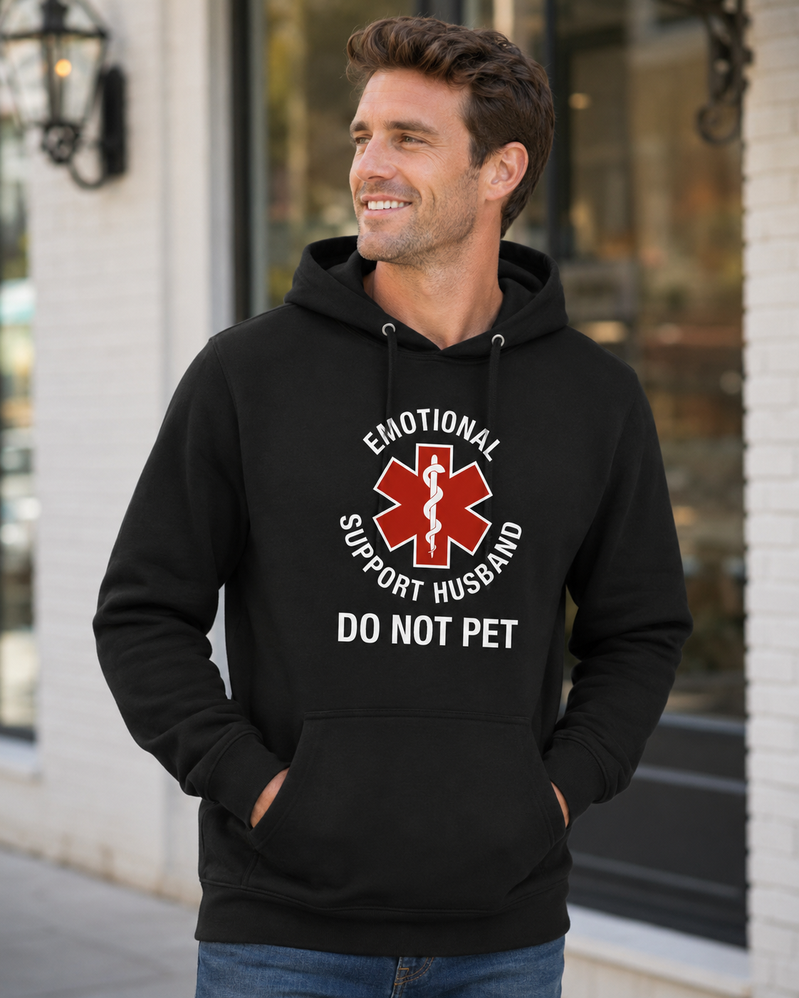 A man wearing a black pullover hoodie with a red and white graphic that reads 'EMOTIONAL SUPPORT HUSBAND DO NOT PET' over a red Star of Life symbol, standing outdoors in front of a storefront.