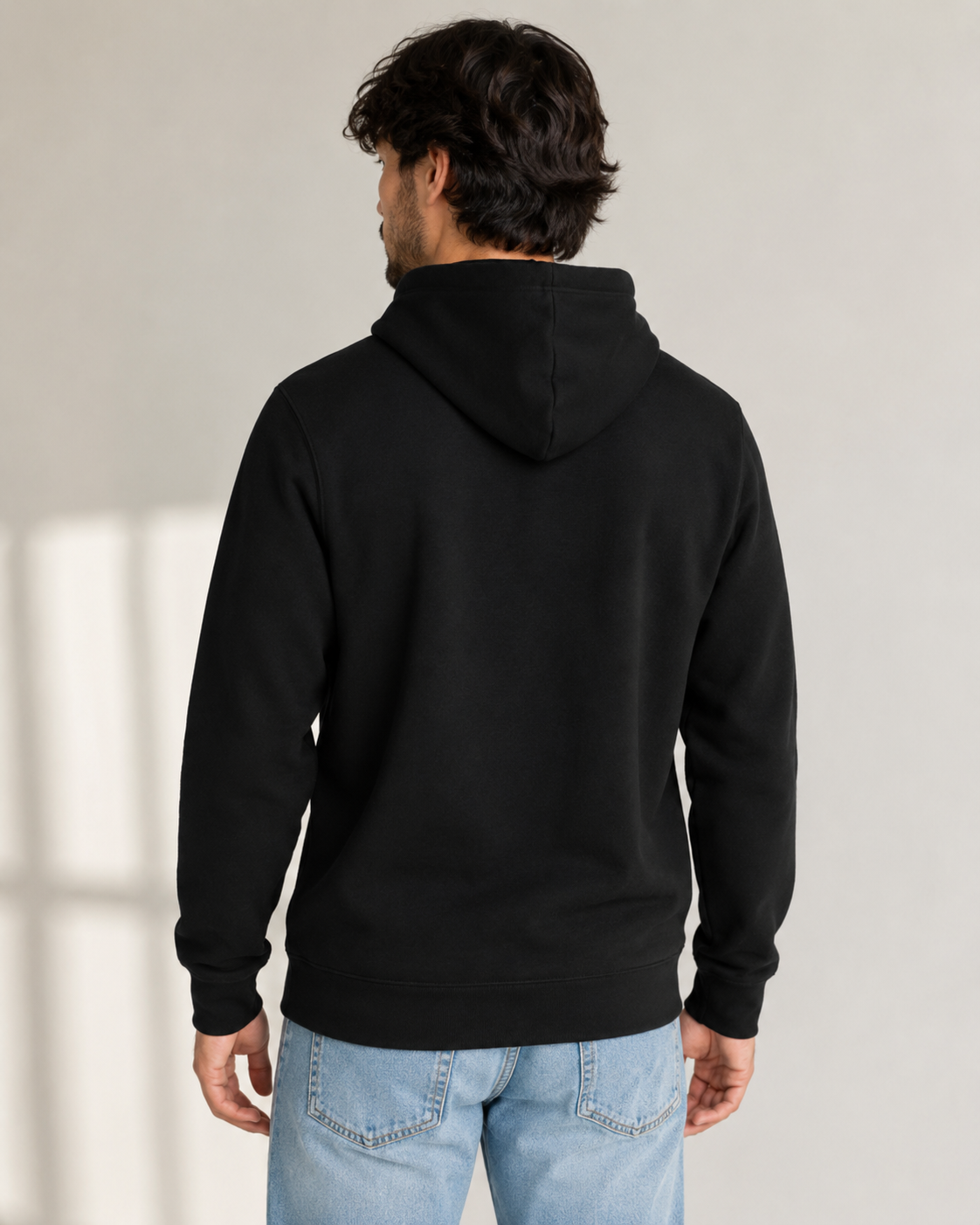 A man viewed from behind wearing a plain black pullover hoodie and light blue jeans, standing against a light neutral wall.