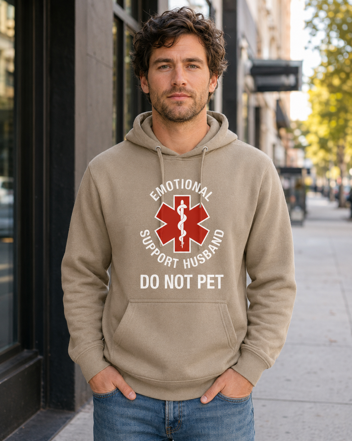A man wearing an olive green hoodie with a red and white graphic that reads 'EMOTIONAL SUPPORT HUSBAND DO NOT PET' in front of a blurred city street background.