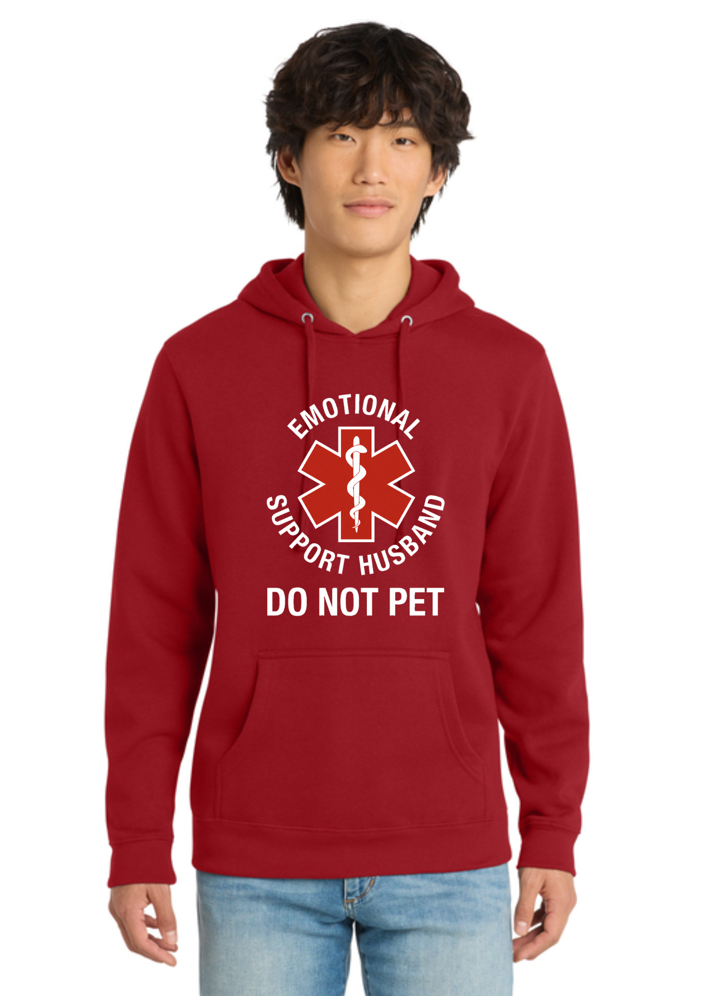 A man wearing a red pullover hoodie with a white graphic print that reads 'EMOTIONAL SUPPORT HUSBAND DO NOT PET' around a red and white star of life symbol. He is also wearing light blue jeans, set against a plain white background.