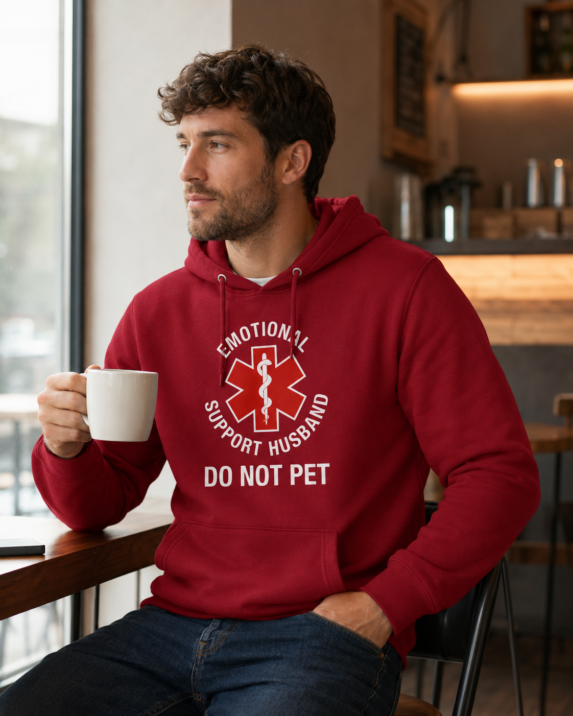 A man in a red hoodie sitting in a cafe, holding a white mug. The hoodie features a white graphic with a red Star of Life and the text 'EMOTIONAL SUPPORT HUSBAND DO NOT PET'.