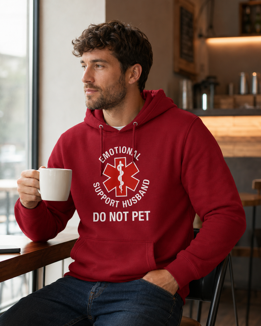 A man in a red hoodie sitting in a cafe, holding a white mug. The hoodie features a white graphic with a red Star of Life and the text 'EMOTIONAL SUPPORT HUSBAND DO NOT PET'.