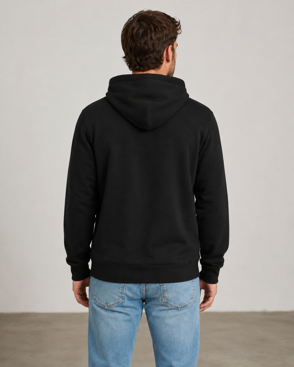 A man viewed from behind wearing a plain black pullover hoodie and light blue jeans, standing against a neutral light gray studio wall.