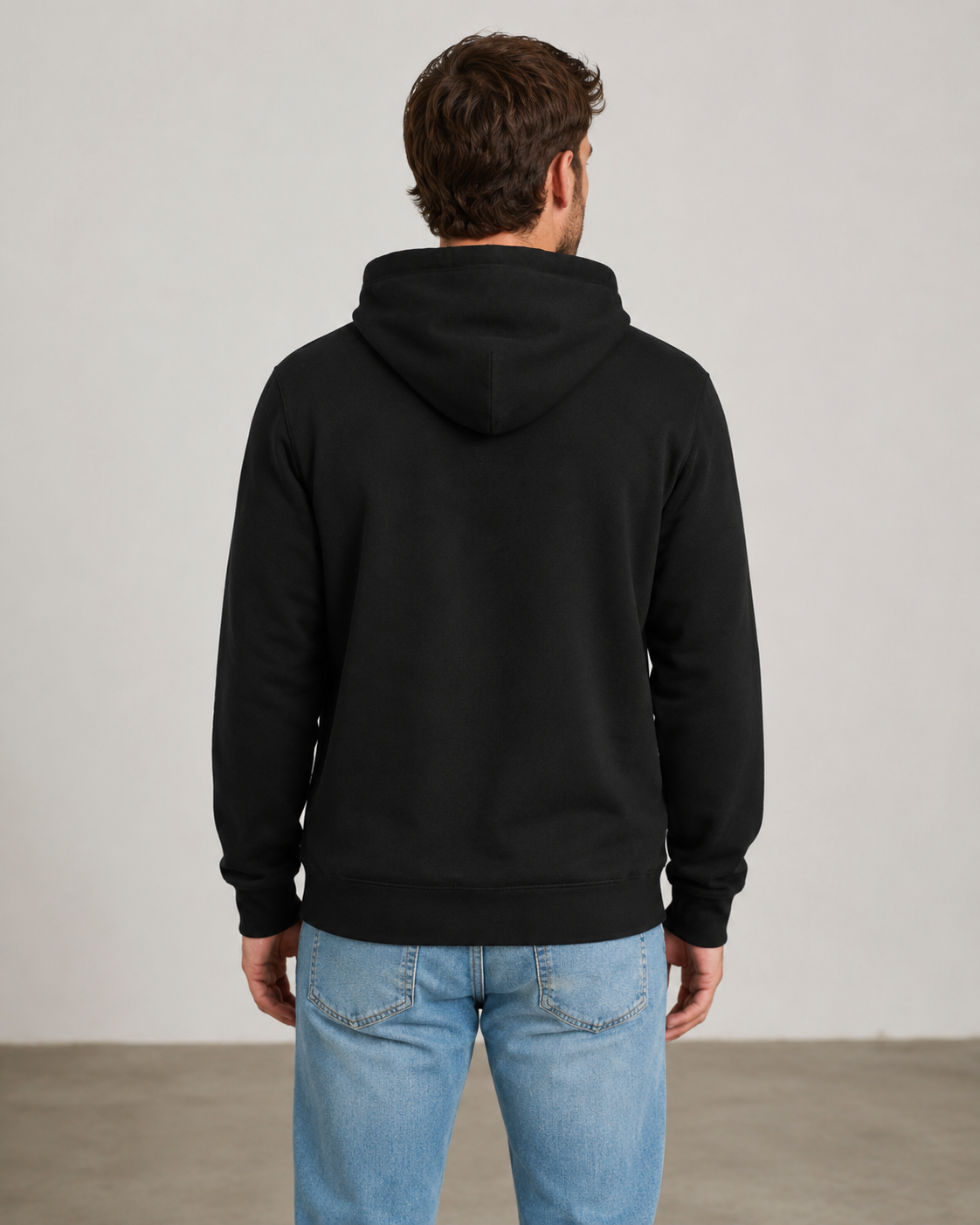A man viewed from behind wearing a plain black pullover hoodie and light blue jeans, standing against a neutral light gray studio wall.