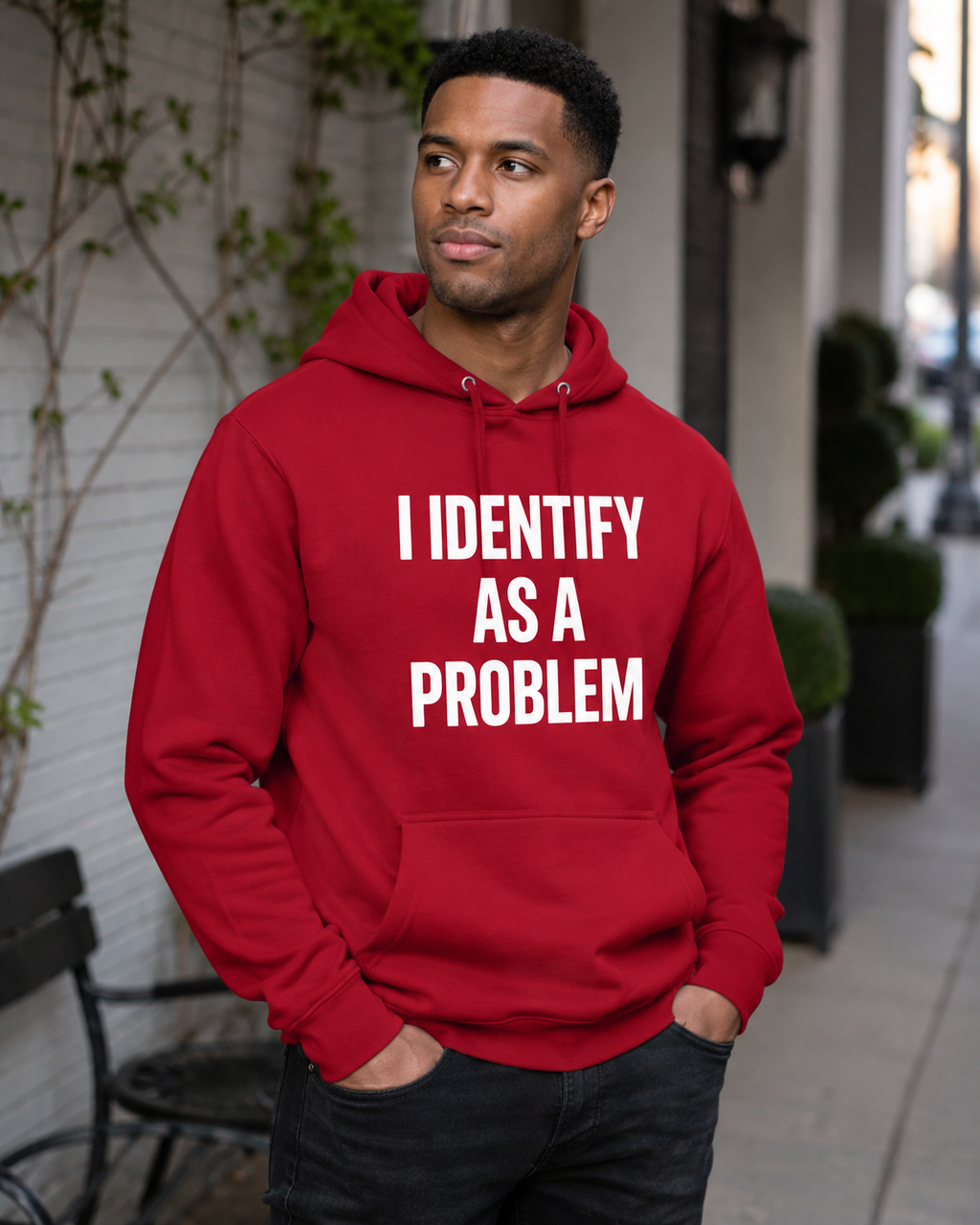 A man wearing a red pullover hoodie with the text 'I IDENTIFY AS A PROBLEM' printed in bold white capital letters on the front, standing outdoors on a sidewalk.