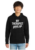 A man wearing a black pullover hoodie with the text 'MY THERAPIST gave up' printed in white on the front, paired with light blue jeans, against a plain white background.