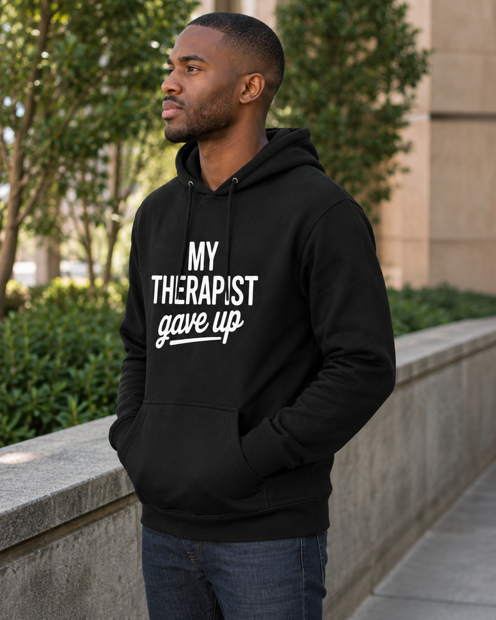 A man wearing a black pullover hoodie with the white text 'MY THERAPIST gave up' printed on the front, standing outdoors next to a stone wall with greenery in the background.