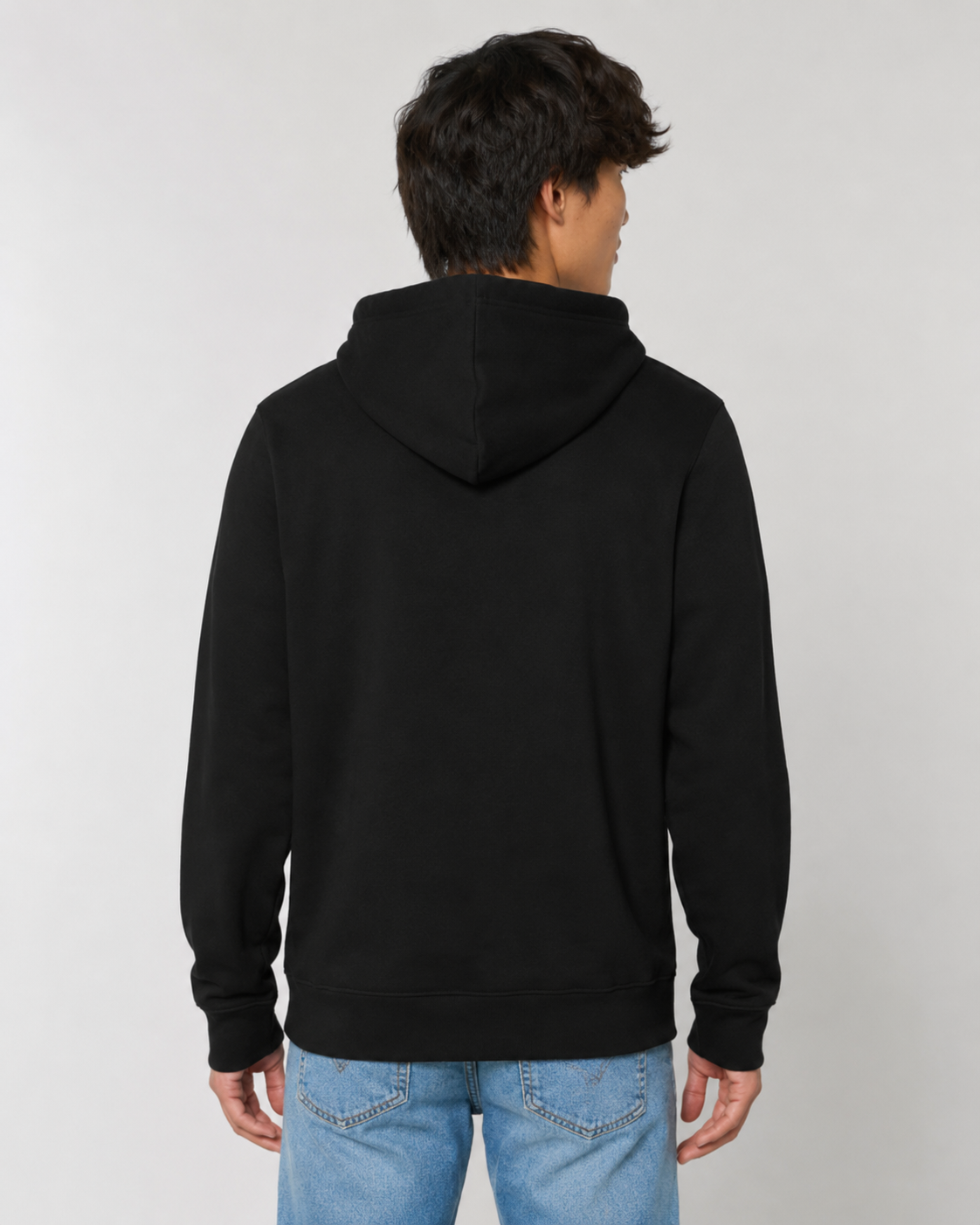 A man viewed from the back wearing a plain black pullover hoodie and light blue jeans, standing against a light gray studio background.
