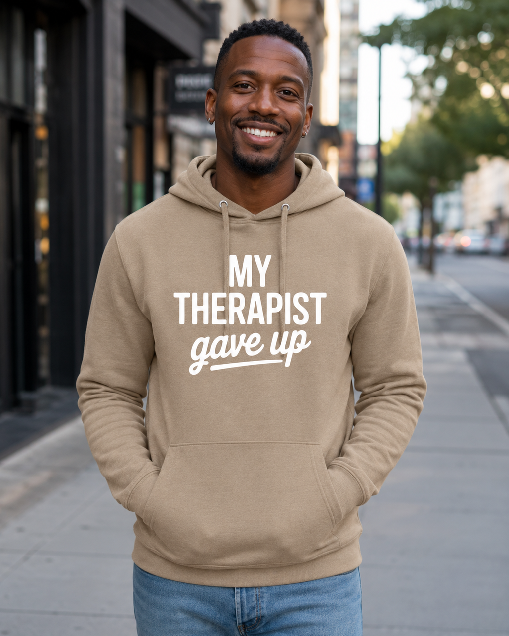 A smiling man wearing an olive green hoodie with the white printed text 'MY THERAPIST gave up' stands on a city sidewalk.