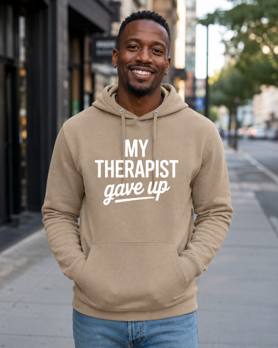 A smiling man wearing an olive green hoodie with the white printed text 'MY THERAPIST gave up' stands on a city sidewalk.