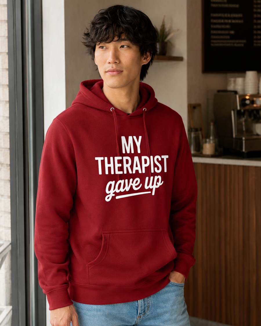 A man wearing a red hoodie with the white printed text 'MY THERAPIST gave up' stands in a cafe-like environment.