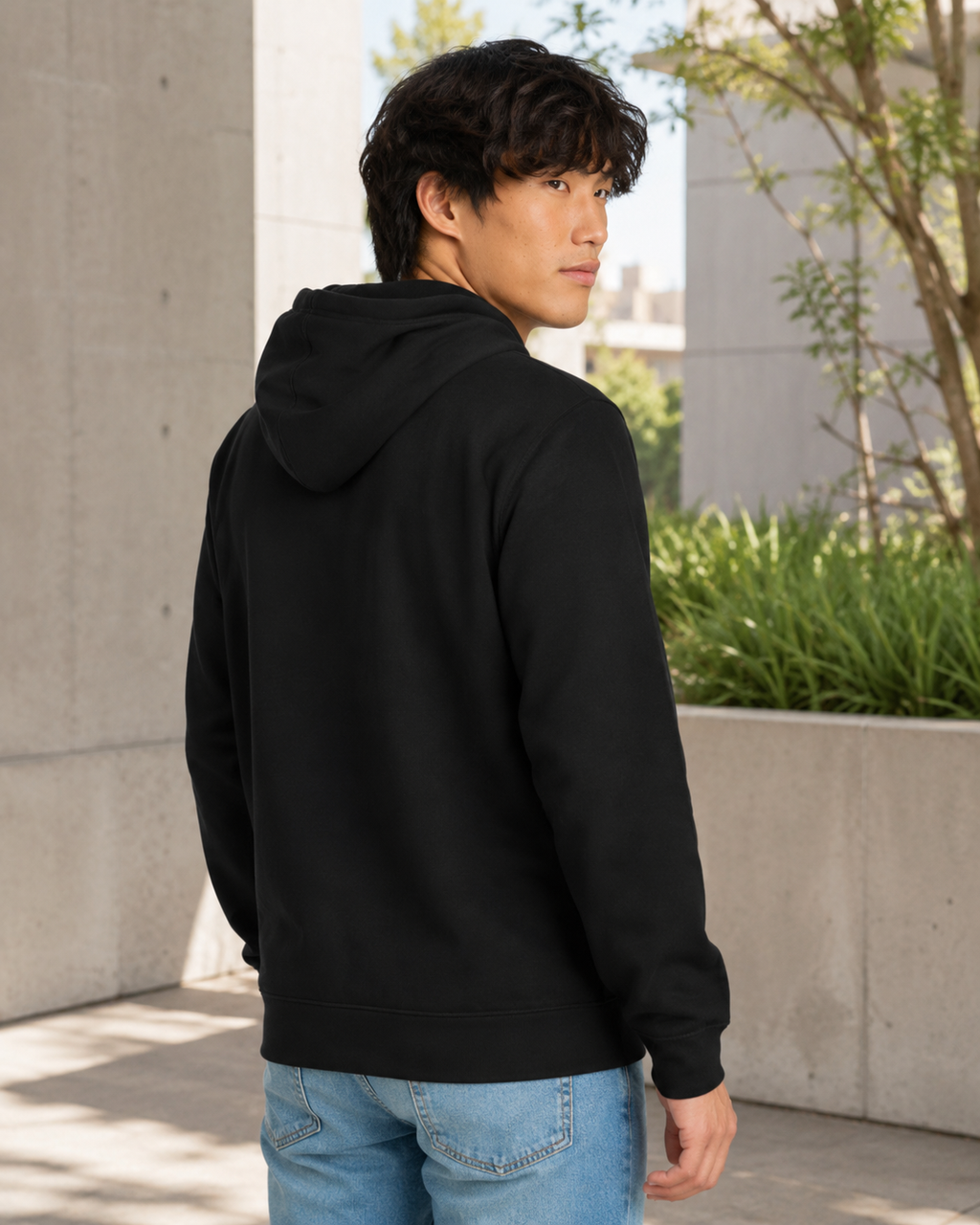 A man wearing a black pullover hoodie and light blue jeans, photographed from the back and side in an outdoor urban setting with concrete walls and greenery.
