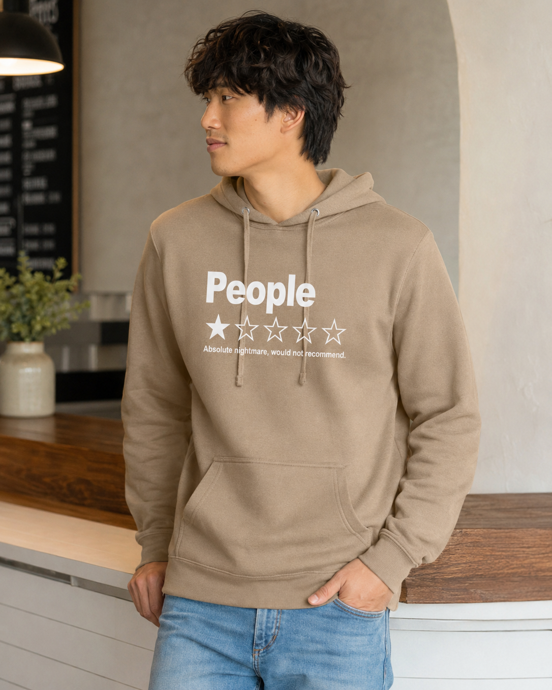 A man wearing an olive green hoodie with a white graphic print that reads 'People' and a five-star rating, paired with blue jeans, standing in a modern indoor setting.