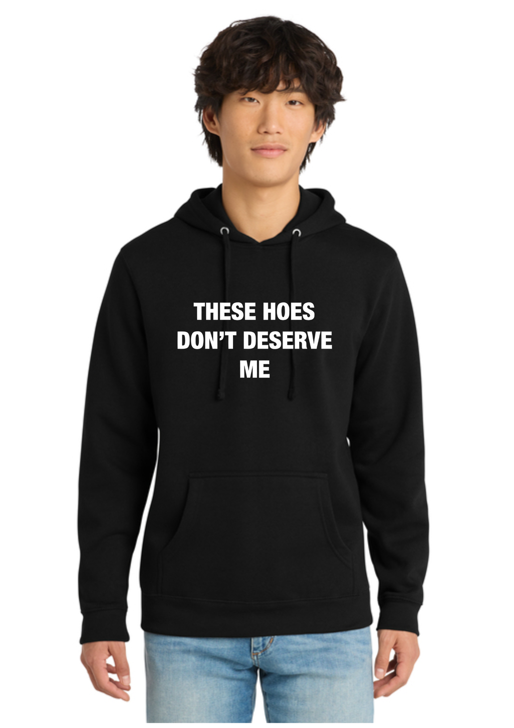 A man wearing a black pullover hoodie with the text 'THESE HOES DON'T DESERVE ME' printed in white capital letters on the chest, paired with light blue jeans, set against a plain white background.
