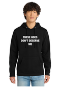 A man wearing a black pullover hoodie with the text 'THESE HOES DON'T DESERVE ME' printed in white capital letters on the chest, paired with light blue jeans, set against a plain white background.