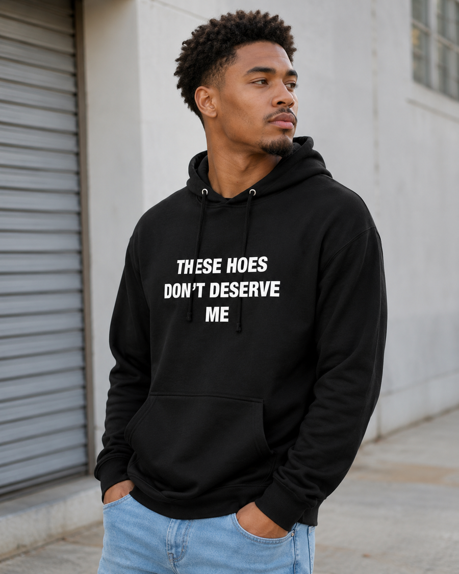 A man wearing a black pullover hoodie with the text 'THESE HOES DON'T DESERVE ME' printed in white capital letters on the chest, paired with light blue jeans, standing outdoors against a light gray building wall.