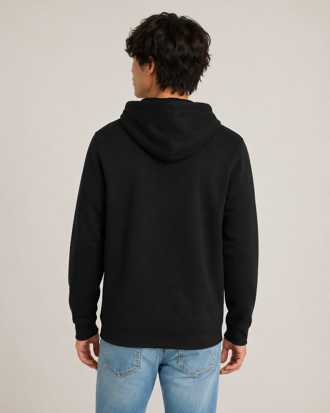 A man viewed from the back wearing a plain black pullover hoodie and light blue jeans, standing against a neutral light gray studio background.
