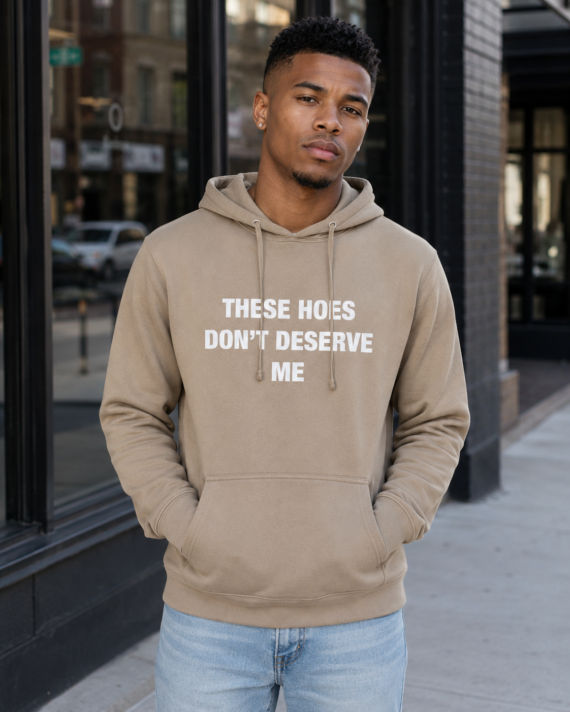 A man wearing an olive green pullover hoodie with the text 'THESE HOES DON'T DESERVE ME' printed in white across the chest, paired with light blue jeans, standing on a city sidewalk.