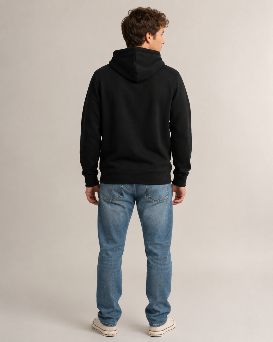 A man viewed from behind wearing a black pullover hoodie, blue jeans, and white sneakers against a plain light gray studio background.