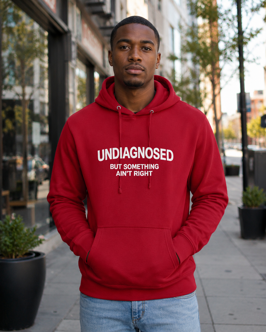 A man wearing a red pullover hoodie with the text 'UNDIAGNOSED BUT SOMETHING AIN'T RIGHT' printed in white on the chest, standing on a city sidewalk.