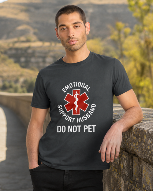 A man wearing a dark gray graphic t-shirt with a red and white 'Emotional Support Husband Do Not Pet' design, standing outdoors against a blurred mountain background.
