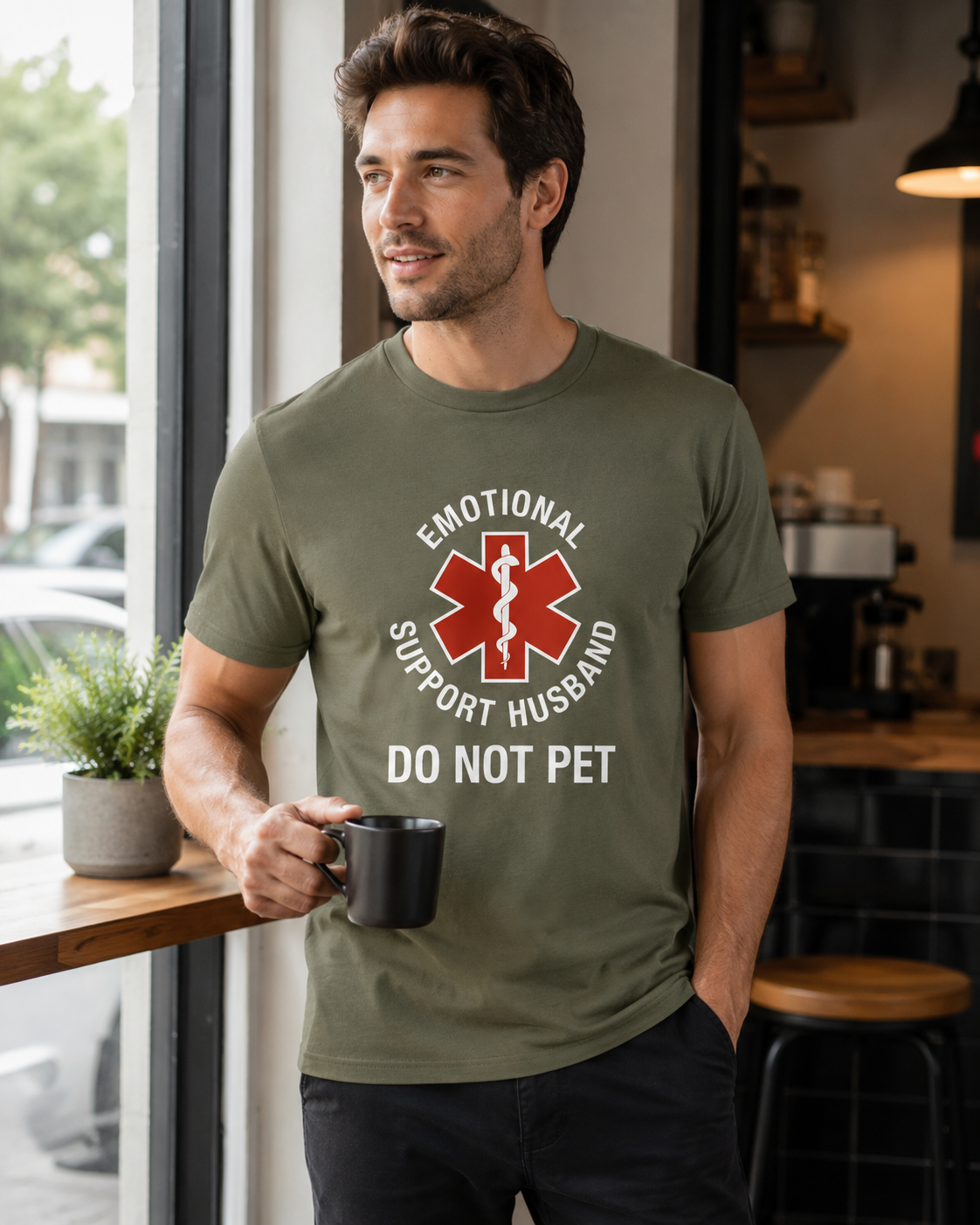 A man wearing an olive green t-shirt with a red and white graphic that reads 'EMOTIONAL SUPPORT HUSBAND DO NOT PET' while holding a black mug in a cafe setting.