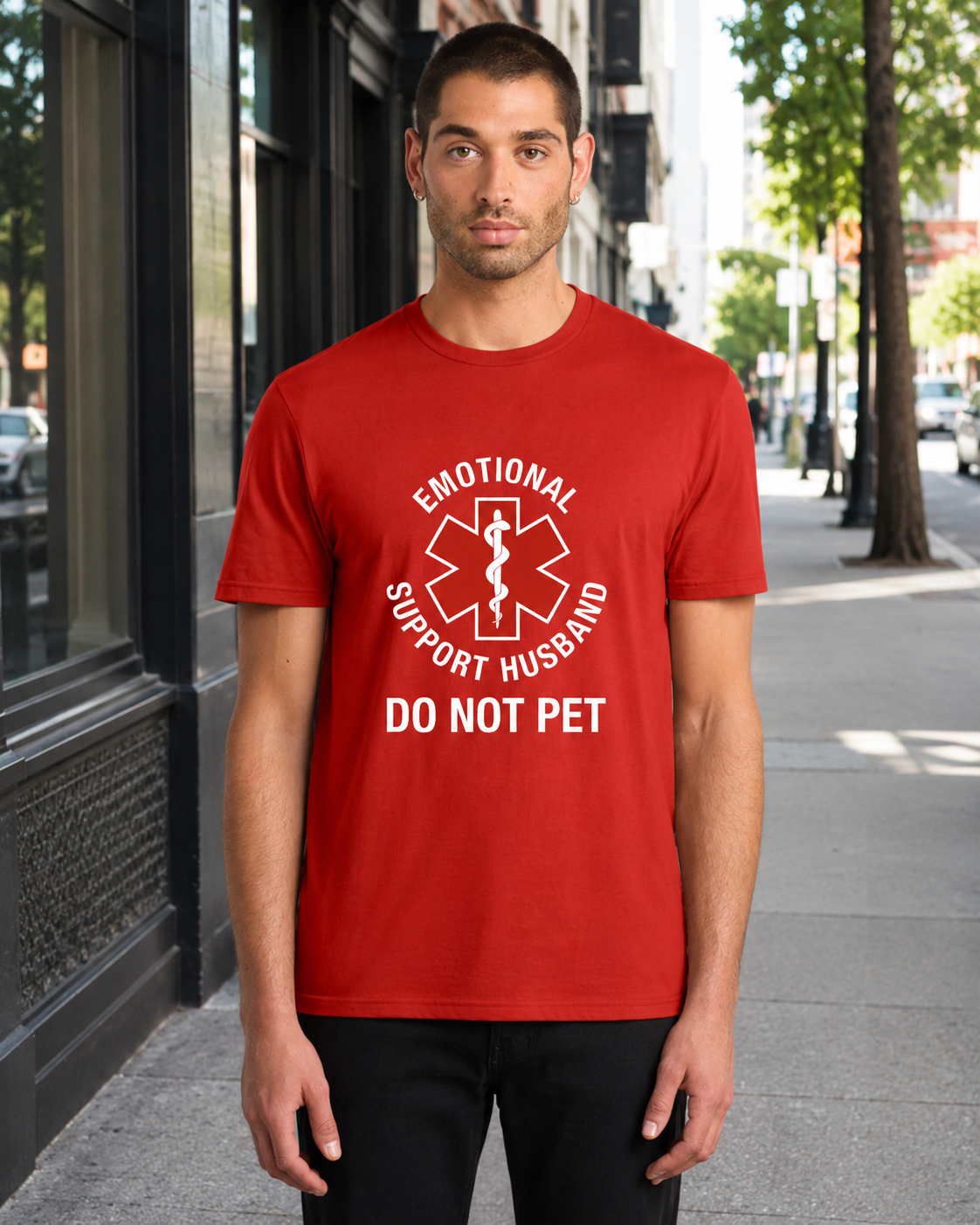 A man wearing a red crewneck t-shirt with a white graphic that reads 'EMOTIONAL SUPPORT HUSBAND DO NOT PET' around a medical-style cross symbol, standing on a city sidewalk.