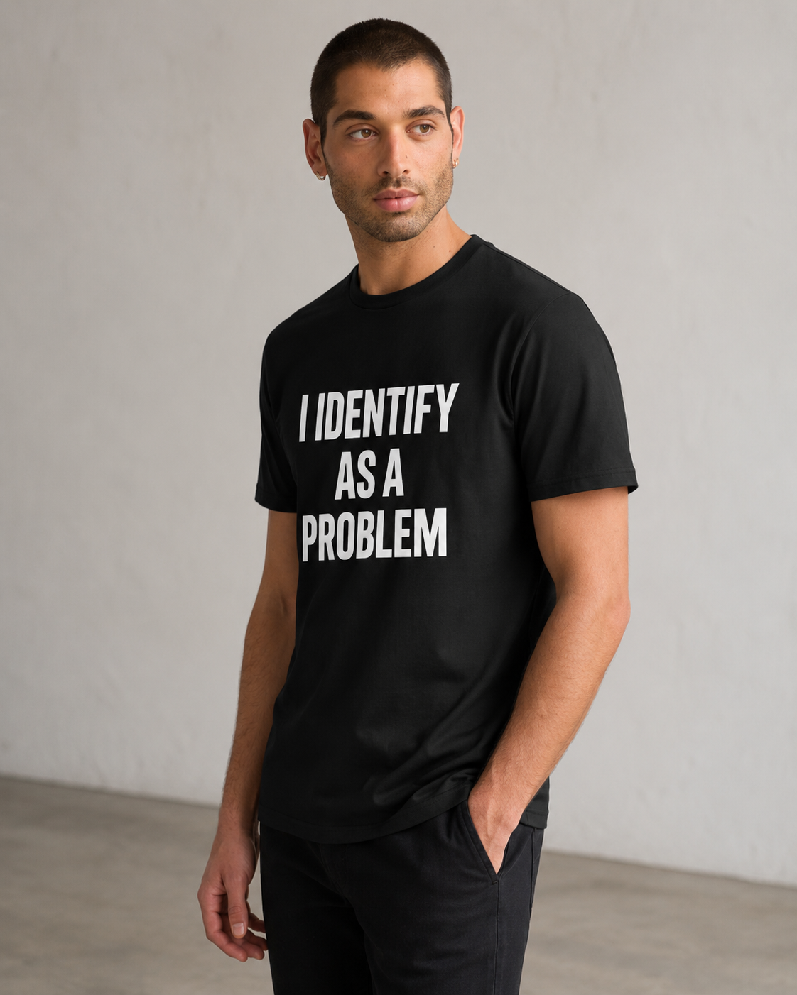 A man wearing a black crewneck t-shirt with the text 'I IDENTIFY AS A PROBLEM' printed in bold white capital letters on the front, standing against a light gray wall.