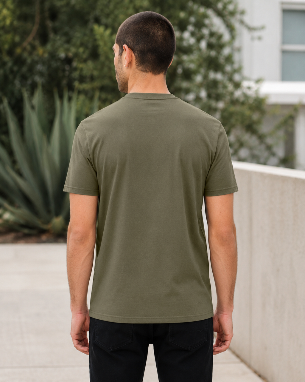 A man viewed from behind wearing a solid olive green short-sleeve t-shirt and black pants, standing outdoors on a paved walkway with a concrete wall and green foliage in the background.