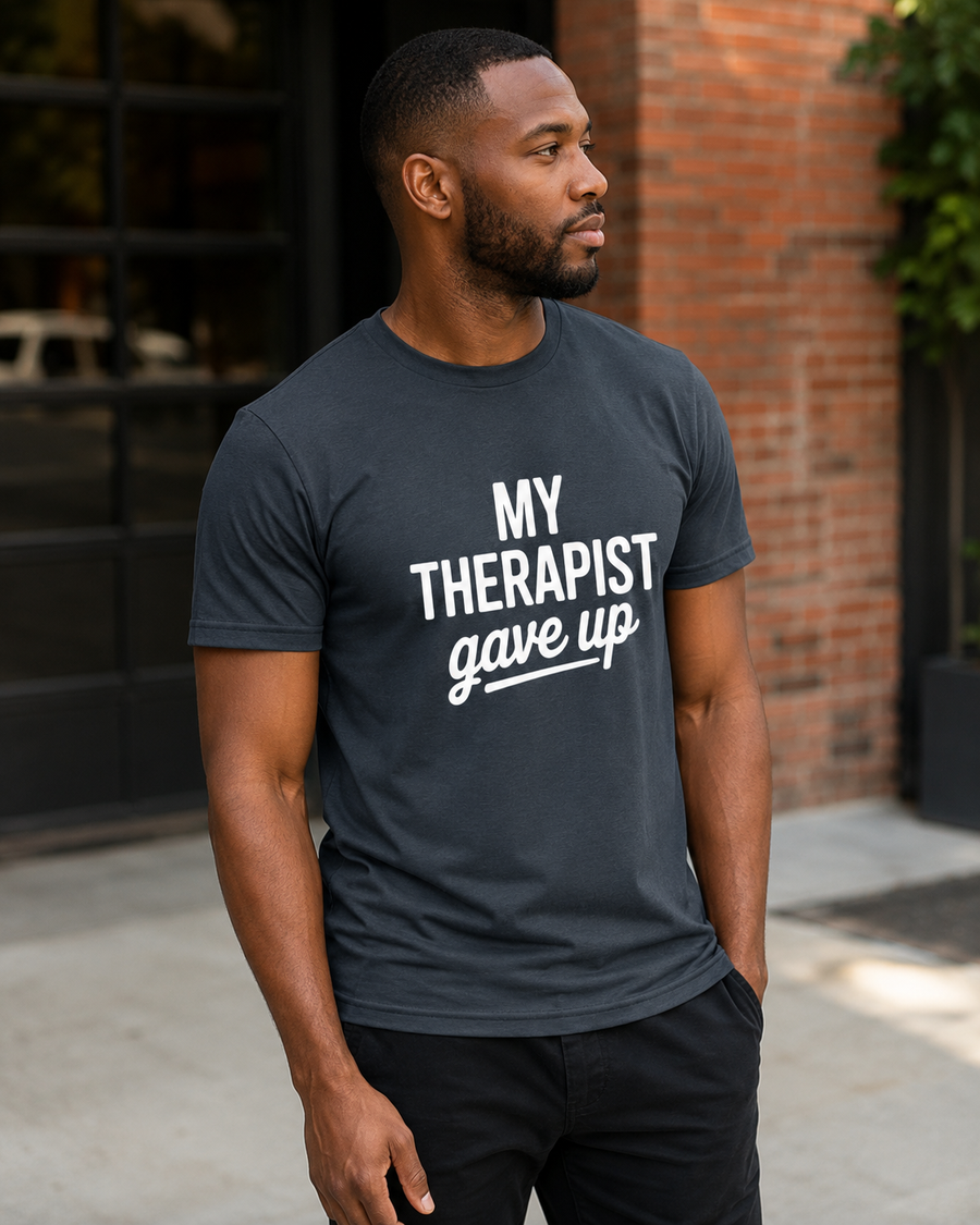 A man wearing a dark gray crewneck t-shirt with the white printed text 'MY THERAPIST gave up' stands outdoors in front of a brick wall and glass door.