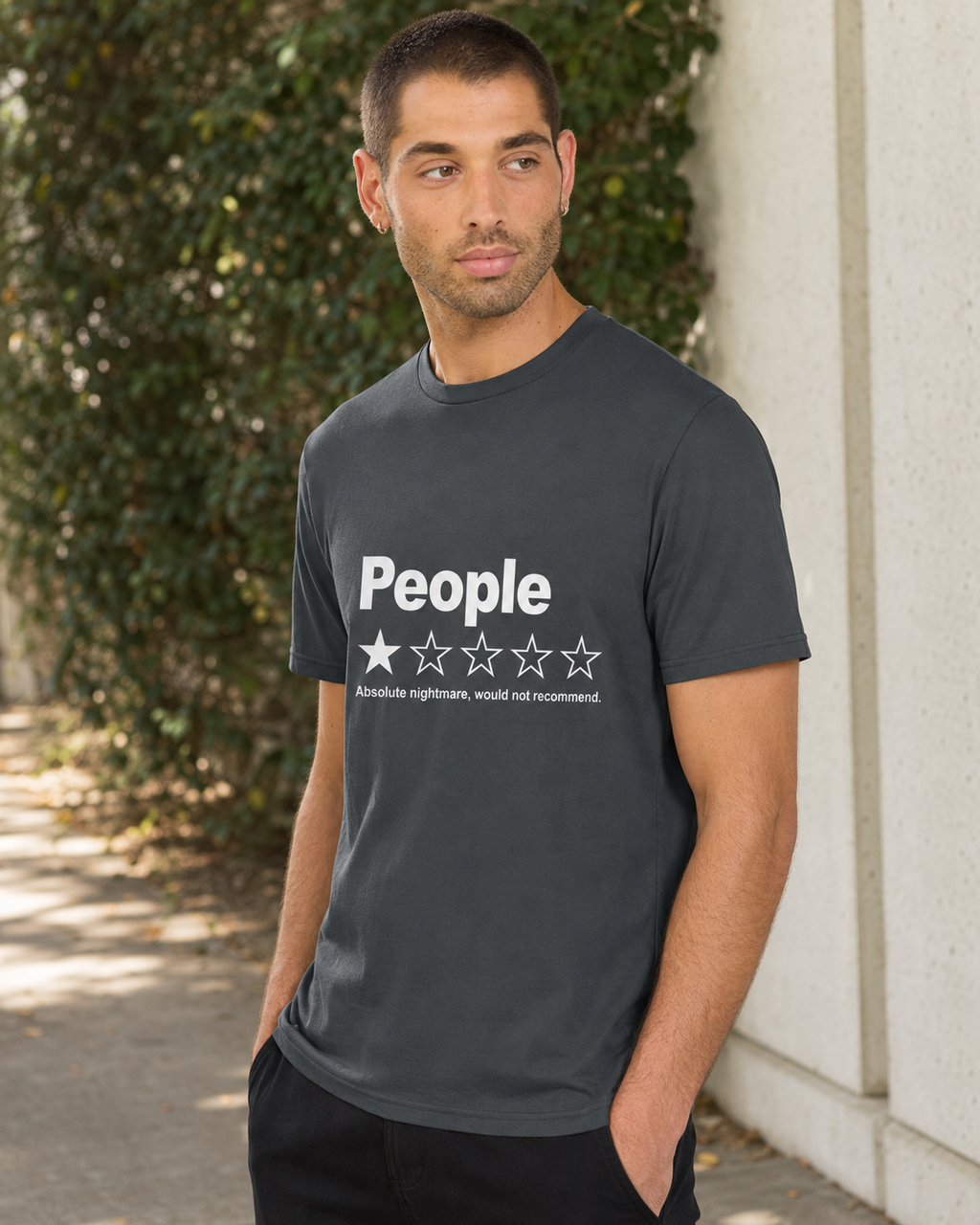A man wearing a dark gray crewneck t-shirt with a white graphic that reads 'People' and a one-star rating, standing outdoors against a concrete wall and green foliage.