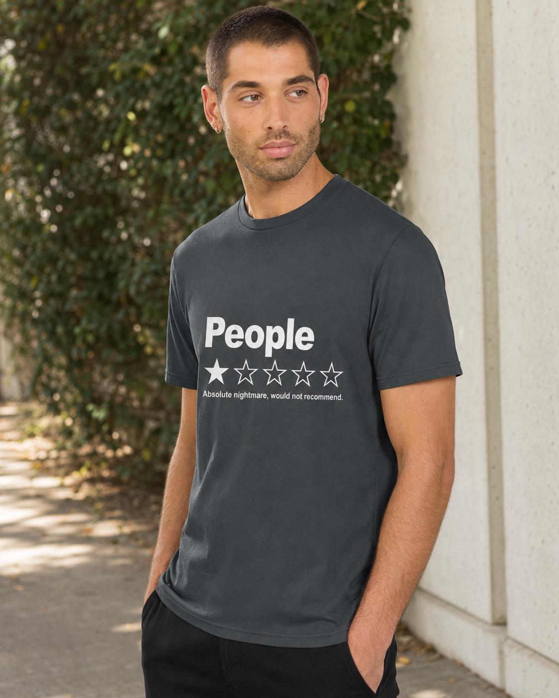 A man wearing a dark gray crewneck t-shirt with a white graphic that reads 'People' and a one-star rating, standing outdoors against a concrete wall and green foliage.