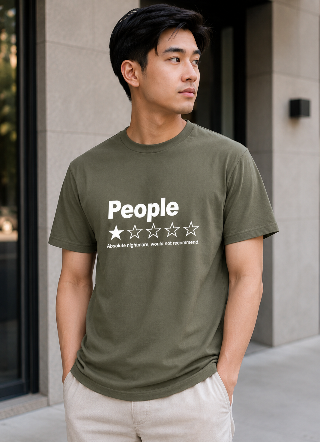 A man wearing an olive green t-shirt with a white graphic print that reads 'People' above a one-star review design, paired with light beige pants, standing outdoors in front of a modern building.