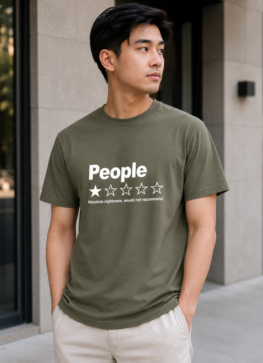 A man wearing an olive green t-shirt with a white graphic print that reads 'People' above a one-star review design, paired with light beige pants, standing outdoors in front of a modern building.