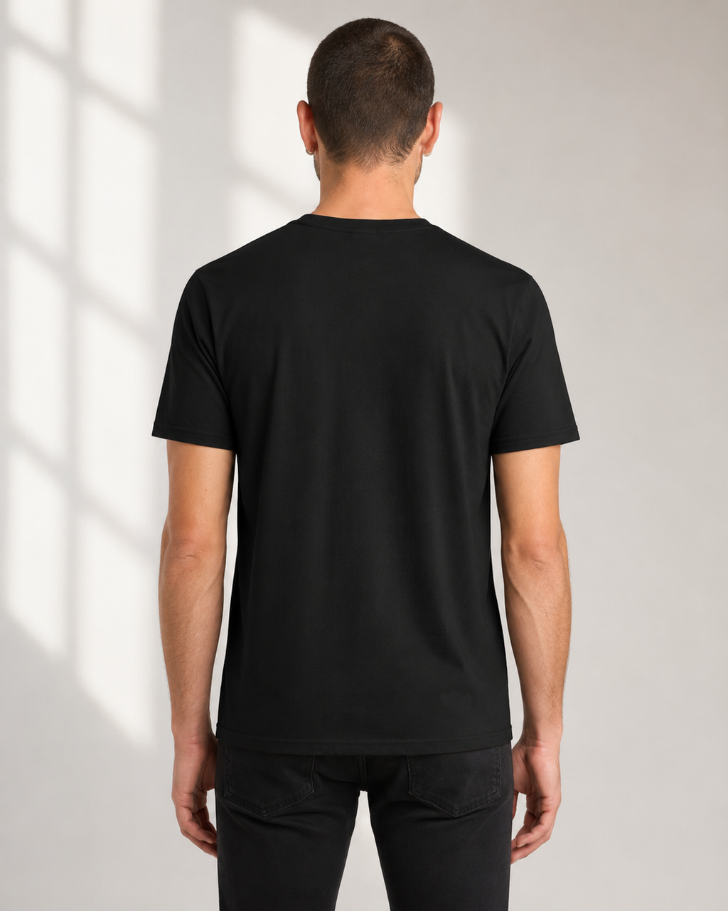 A man viewed from behind wearing a plain black short-sleeve crew-neck t-shirt and black pants, standing against a light gray wall with soft window shadow patterns.
