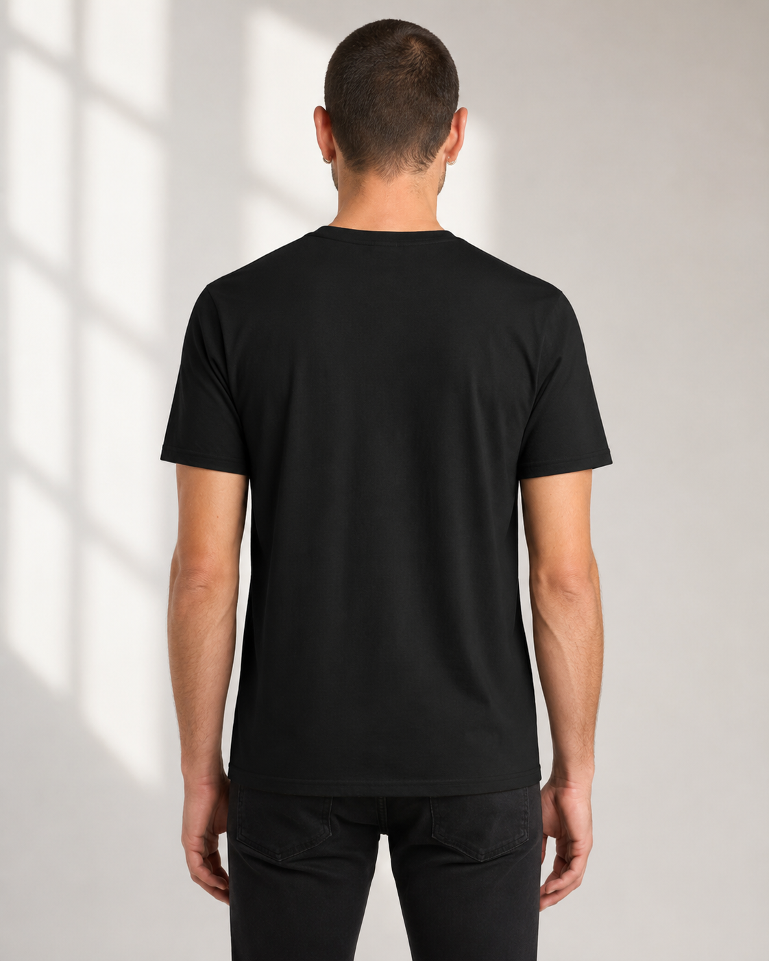 A man viewed from behind wearing a plain black short-sleeve crew-neck t-shirt and black pants, standing against a light gray wall with soft window shadow patterns.