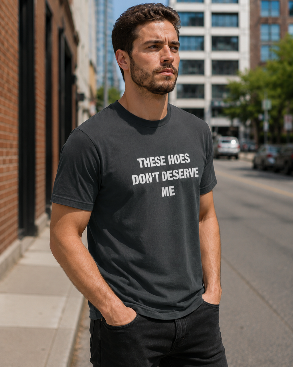 A man wearing a dark charcoal gray t-shirt with the white printed text 'THESE HOES DON'T DESERVE ME' standing on a city street.