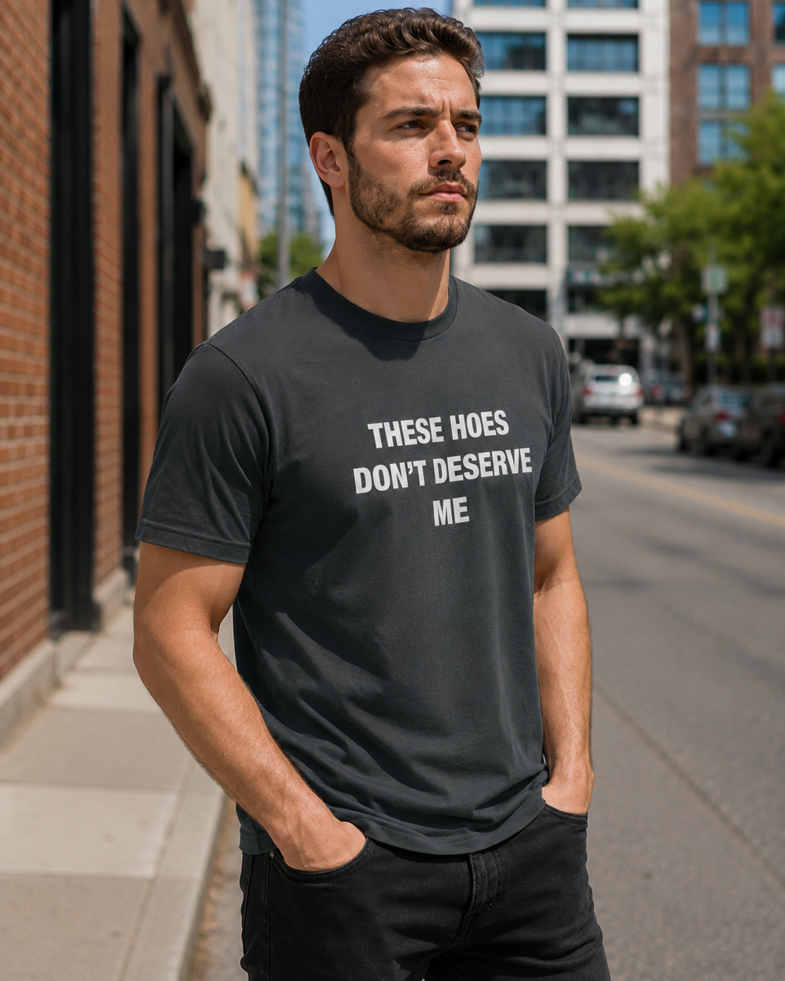 A man wearing a dark charcoal gray t-shirt with the white printed text 'THESE HOES DON'T DESERVE ME' standing on a city street.