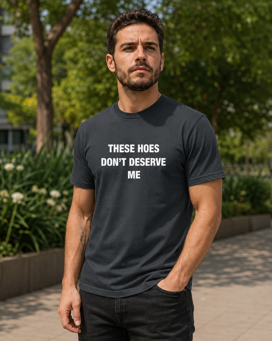 A man wearing a dark gray graphic t-shirt with the text 'THESE HOES DON'T DESERVE ME' in white, standing outdoors on a paved path with trees in the background.