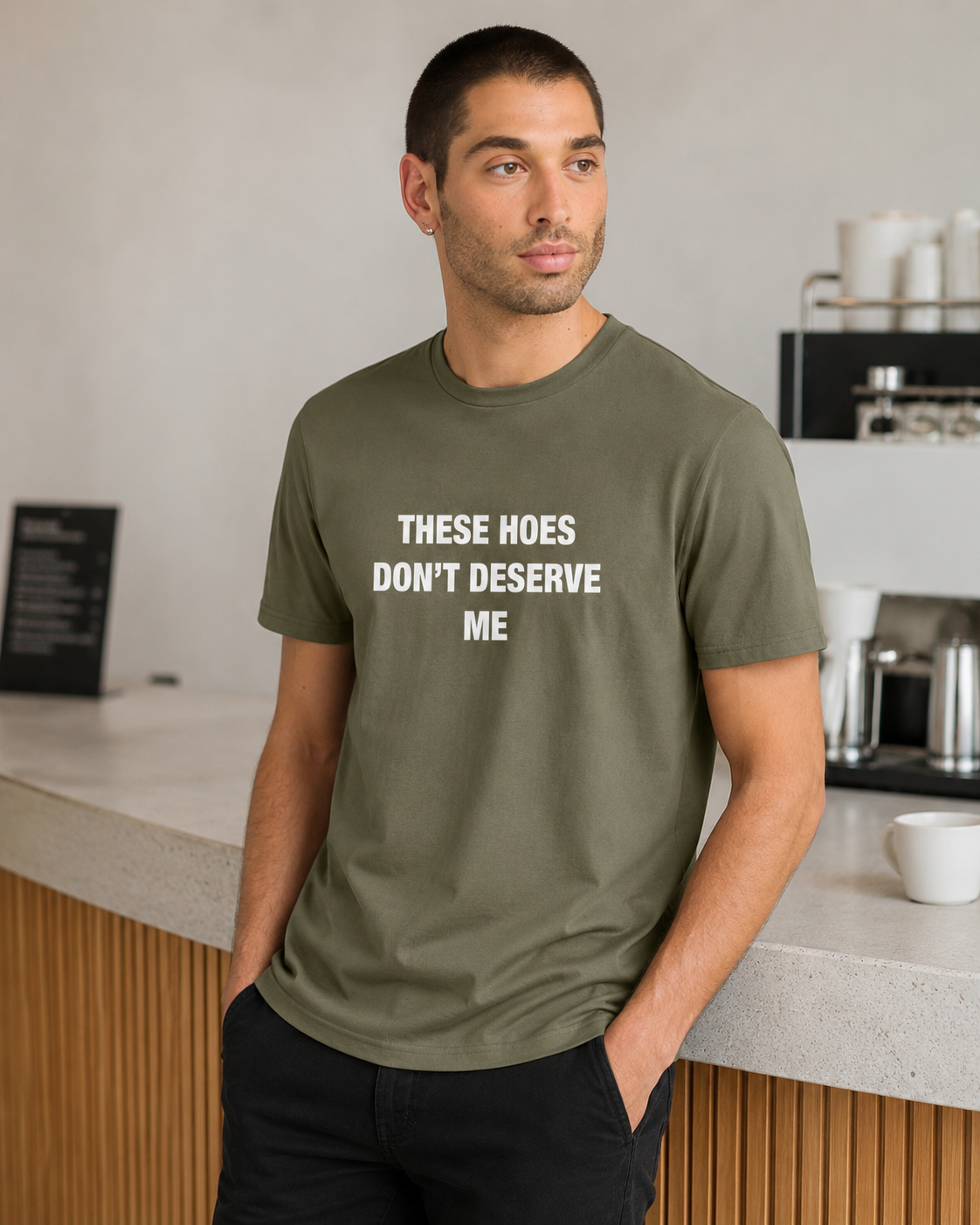 A man wearing an olive green t-shirt with the white printed text 'THESE HOES DON'T DESERVE ME' standing in a modern cafe setting.