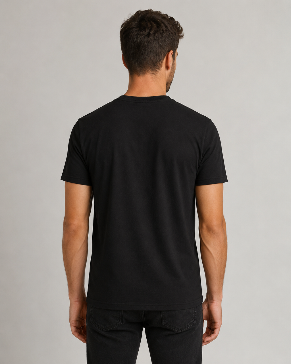 A man viewed from the back wearing a plain black short-sleeve crewneck t-shirt and black jeans, standing against a neutral light gray studio background.