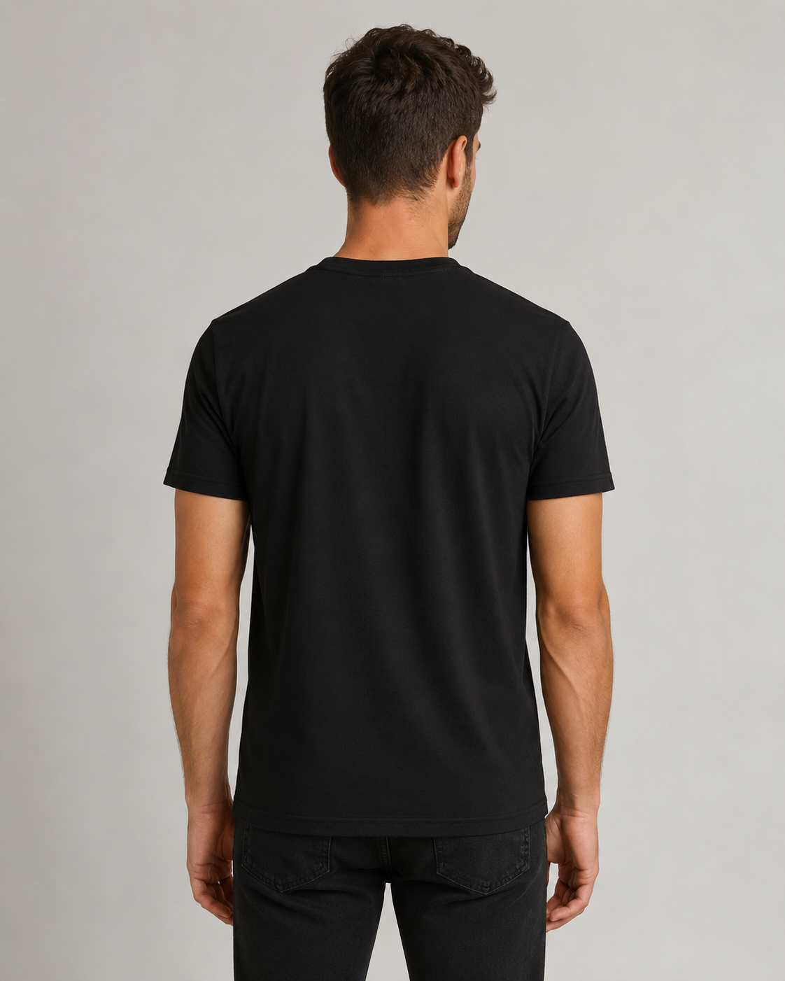 A man viewed from the back wearing a plain black short-sleeve crewneck t-shirt and black jeans, standing against a neutral light gray studio background.