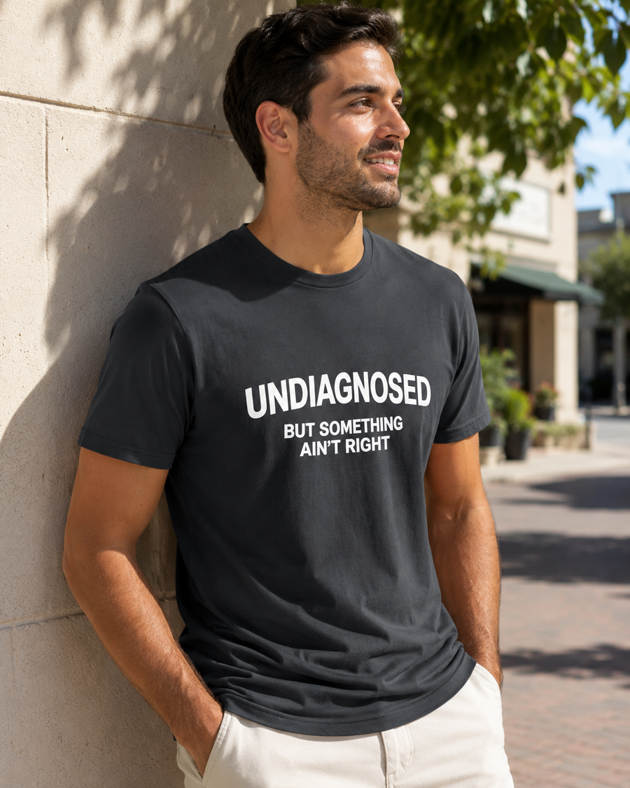 A man wearing a dark charcoal gray graphic t-shirt with white text that reads 'UNDIAGNOSED BUT SOMETHING AIN'T RIGHT', leaning against a light-colored stone wall in an outdoor setting.